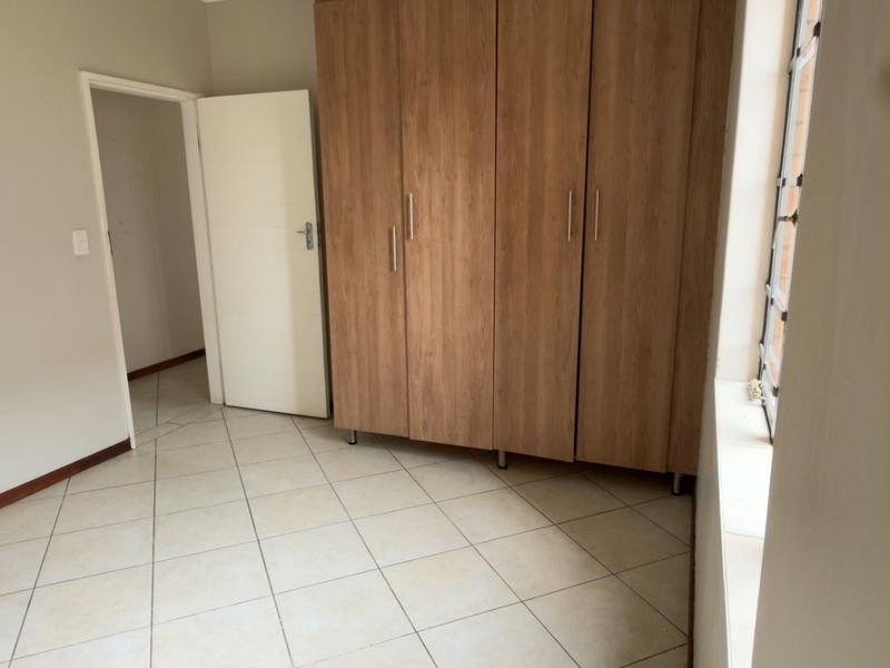 A bright, empty bedroom featuring neutral tiled flooring, built-in wooden cupboards, and a window with security bars.