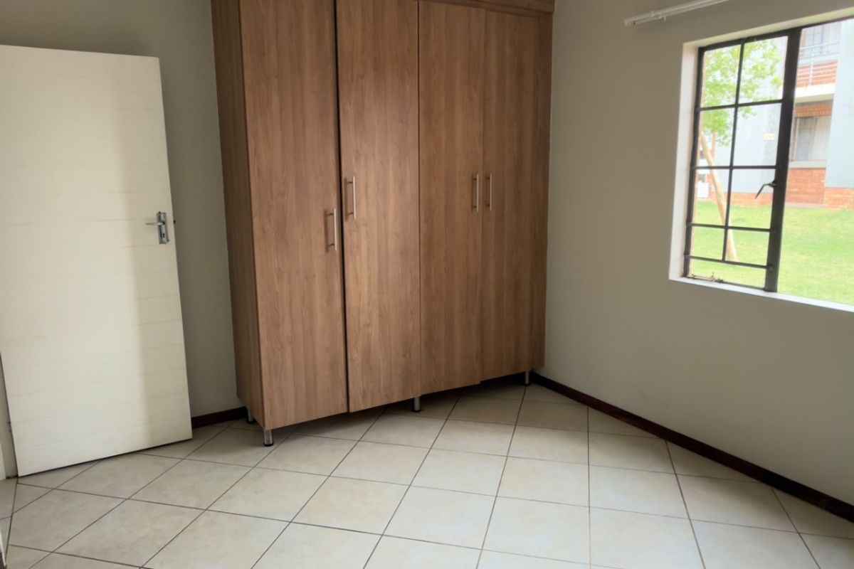 A clean, empty bedroom featuring neutral tiled flooring, a built-in wooden wardrobe, and a window with security bars overlooking the exterior grounds.