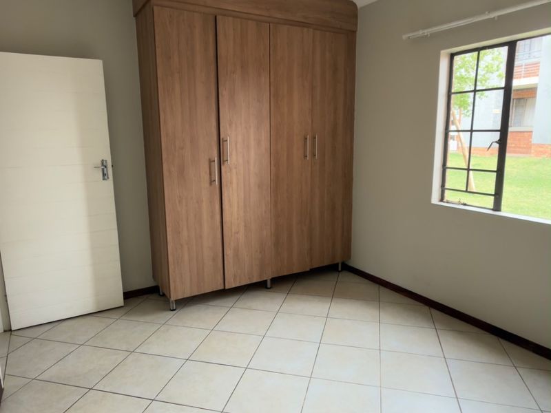 A clean, empty bedroom featuring neutral tiled flooring, a built-in wooden wardrobe, and a window with security bars overlooking the exterior grounds.