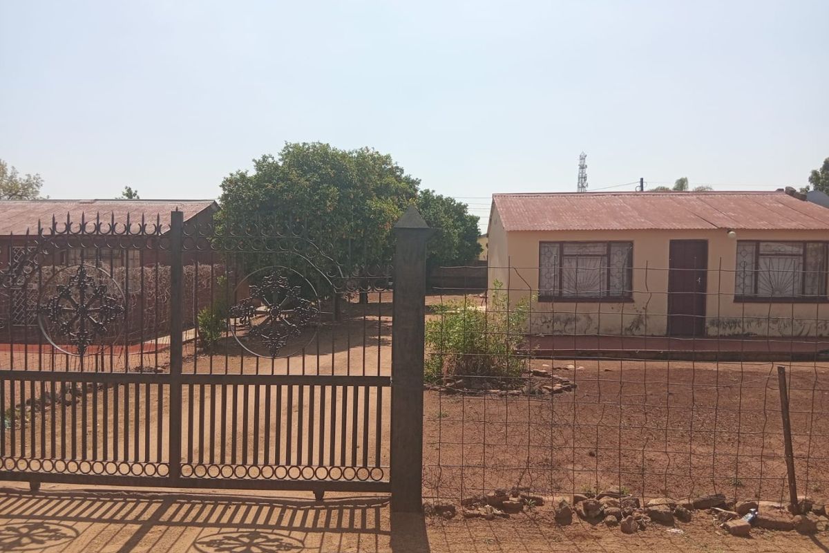 Front view of a residential property featuring a metal security gate, wire fencing, and a house with a corrugated metal roof and security bars.
