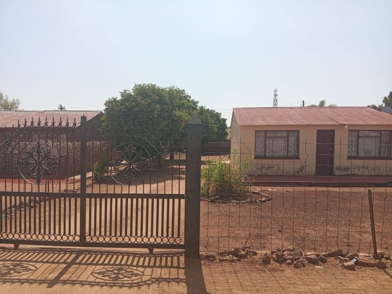 Front view of a residential property featuring a metal security gate, wire fencing, and a house with a corrugated metal roof and security bars.