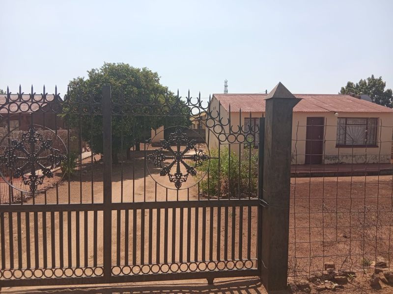Front view of a residential property featuring a decorative wrought iron gate, wire fencing, and a single-story house with a metal roof.