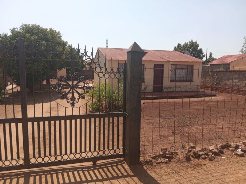 Front view of a single-story house featuring a corrugated metal roof, security bars on windows, and a gated entrance with an unpaved yard.