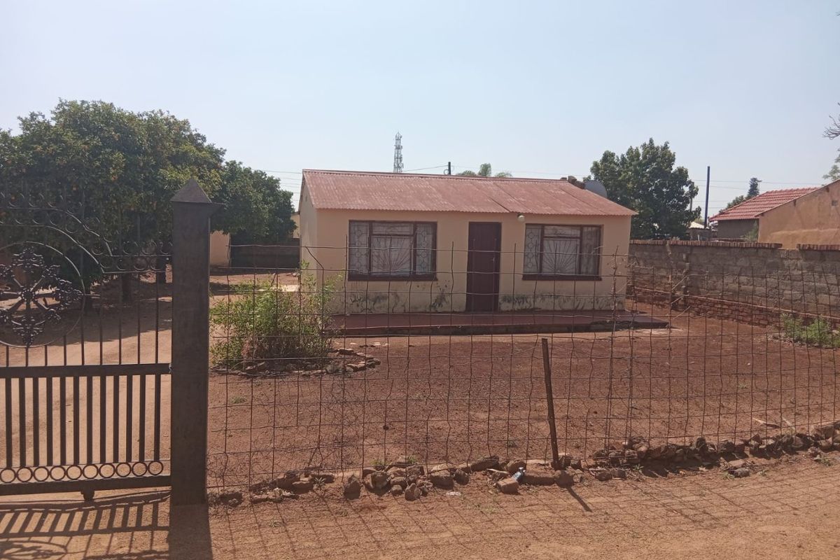 Front view of a detached house featuring a corrugated metal roof, concrete porch, and perimeter fencing on a large plot.