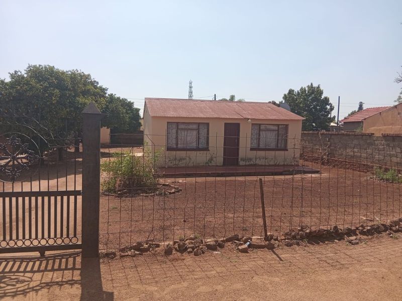Front view of a detached house featuring a corrugated metal roof, concrete porch, and perimeter fencing on a large plot.