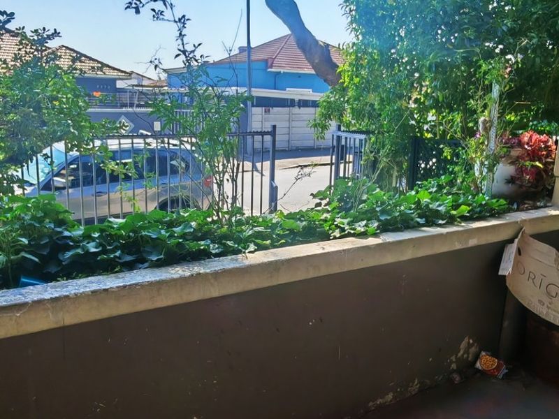 A covered balcony area featuring a concrete balustrade and flooring, showing signs of wear and weathering, overlooking a street-facing garden area.