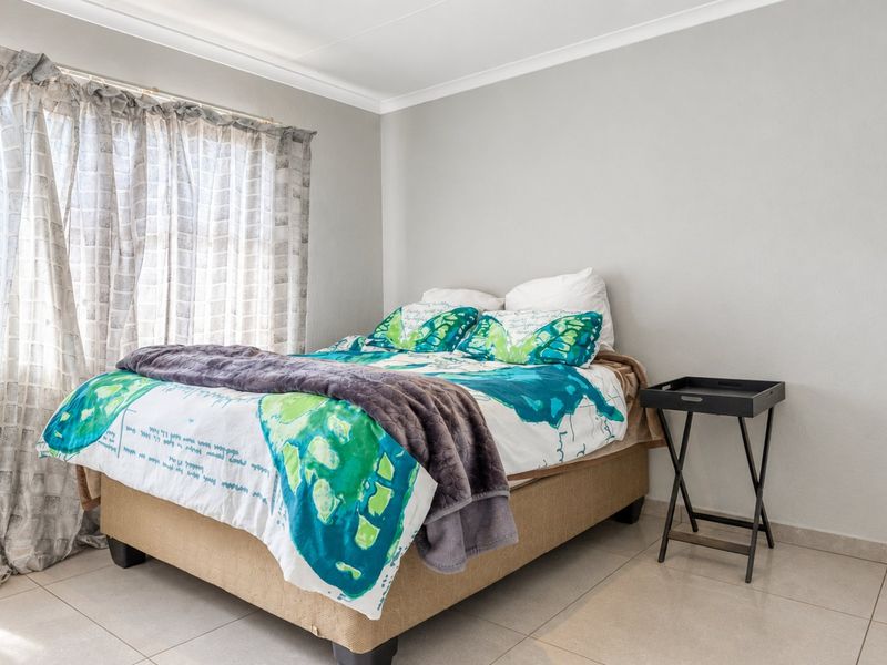 A well-maintained bedroom featuring neutral tiled flooring, clean wall finishes, and natural light provided by a large window with curtain rails.