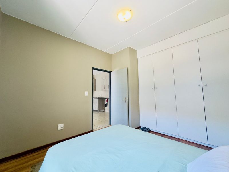 A well-maintained bedroom featuring built-in cupboards, laminate flooring, and a neutral color palette, with a doorway leading to an adjacent kitchen area.