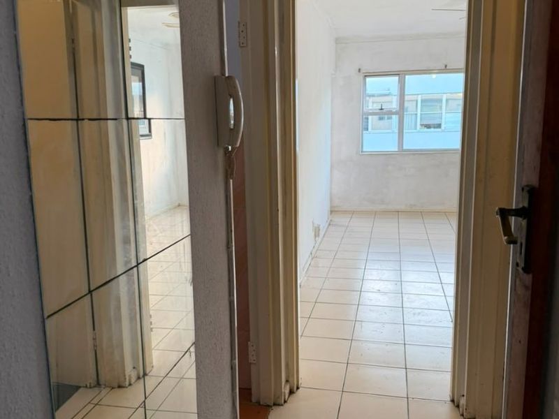 A vacant hallway featuring tiled flooring, an intercom system, and a wall-mounted mirror, leading into an adjacent room with natural light.