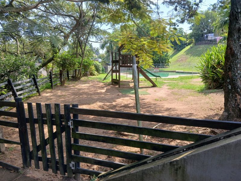 A communal outdoor area featuring a swimming pool, playground equipment, and perimeter fencing, set within a landscaped garden environment.