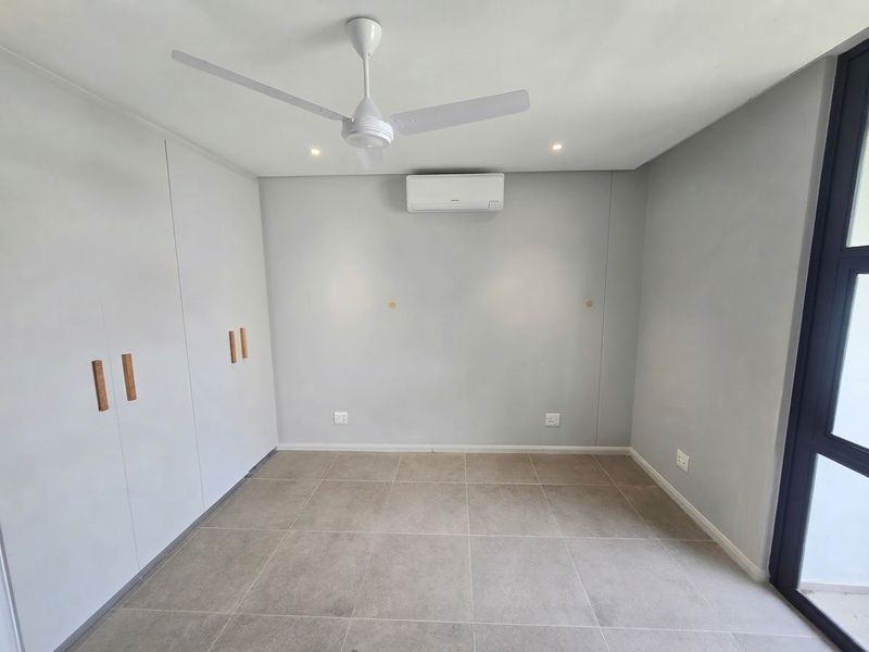A clean, modern bedroom featuring neutral tiled flooring, built-in cupboards, a ceiling fan, and a wall-mounted air conditioning unit.