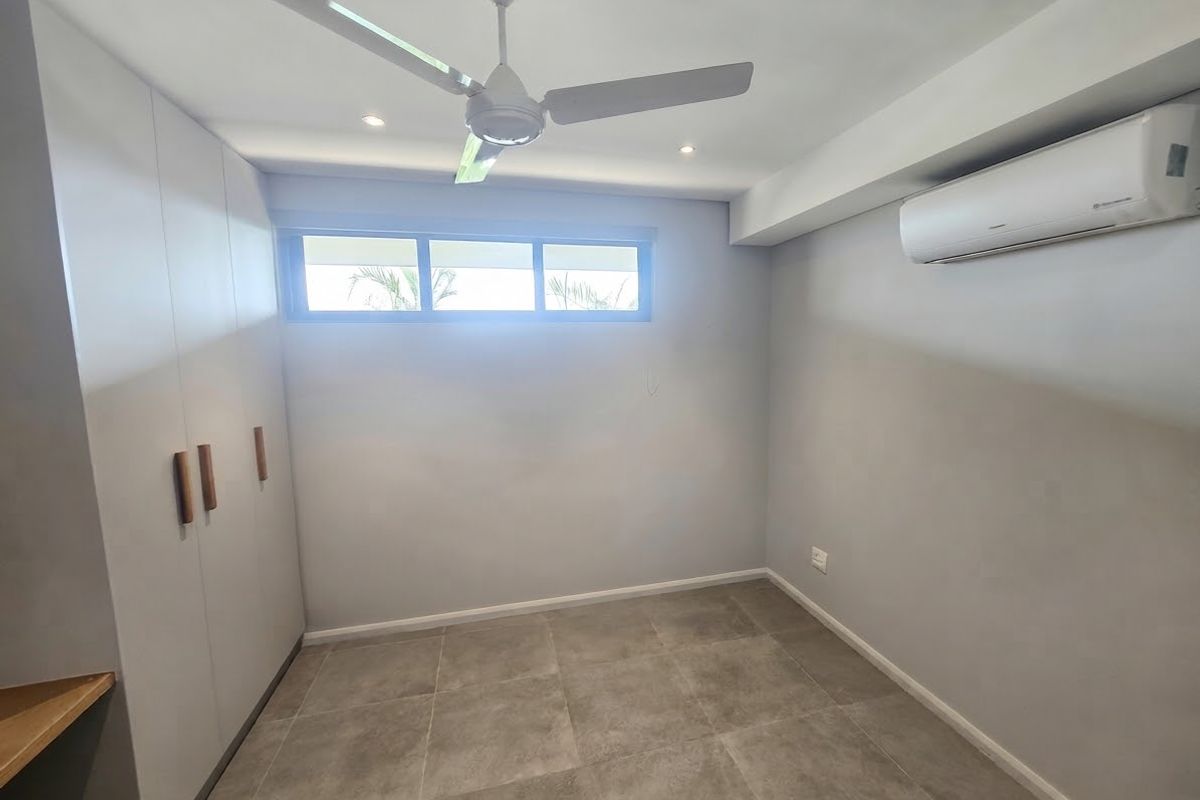 A clean, modern bedroom featuring neutral tiled flooring, built-in cupboards, a ceiling fan, and a wall-mounted split-system air conditioner.