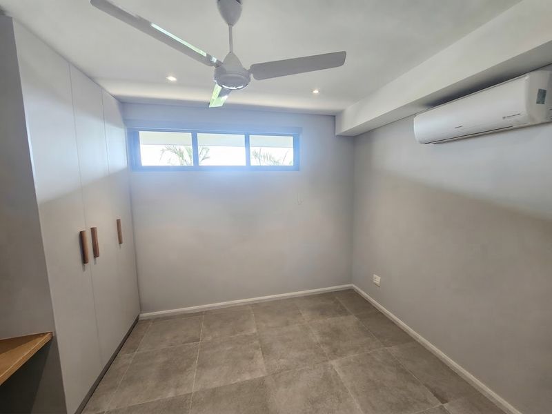 A clean, modern bedroom featuring neutral tiled flooring, built-in cupboards, a ceiling fan, and a wall-mounted split-system air conditioner.