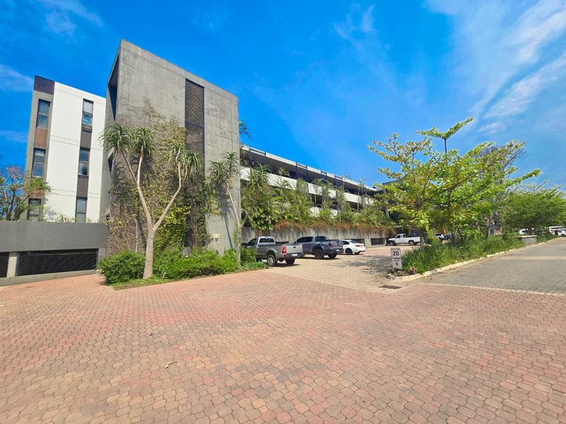 Exterior view of a modern apartment complex featuring a paved driveway, concrete facade, and integrated multi-level parking structure with landscaped surroundings.