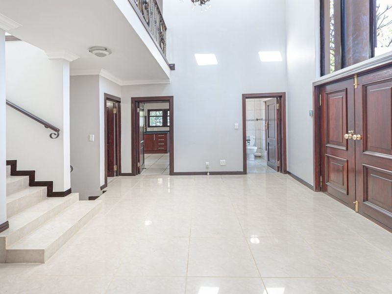 Spacious double-volume entrance hall featuring tiled flooring, a grand wooden staircase with wrought iron balustrade, and natural light from overhead skylights.