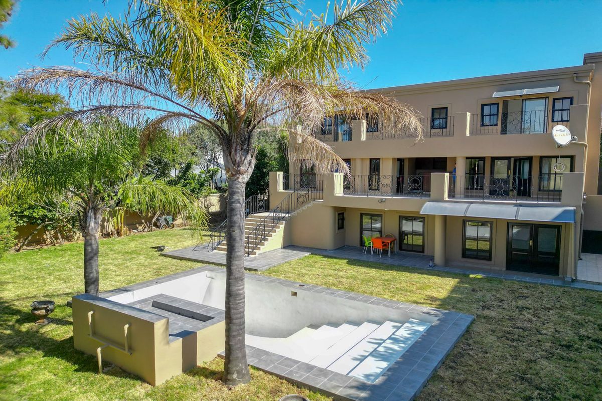 Rear view of a multi-story house featuring a swimming pool, landscaped garden, elevated balcony with wrought iron railings, and a paved patio area.