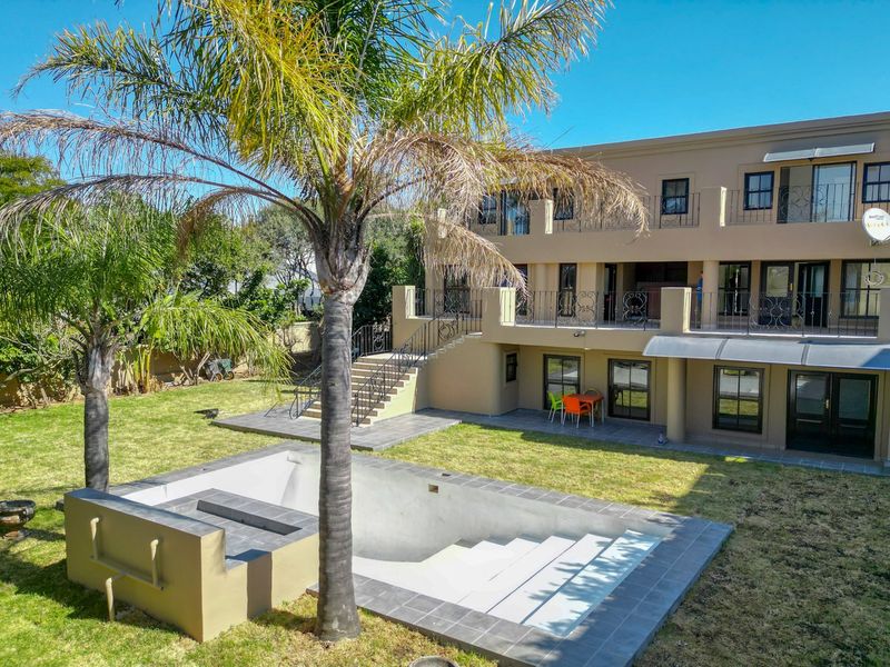 Rear view of a multi-story house featuring a swimming pool, landscaped garden, elevated balcony with wrought iron railings, and a paved patio area.