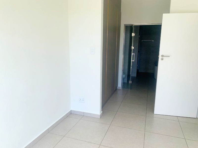 A clean, empty bedroom featuring neutral tiled flooring, built-in cupboards, and a glass door leading to an en-suite bathroom.