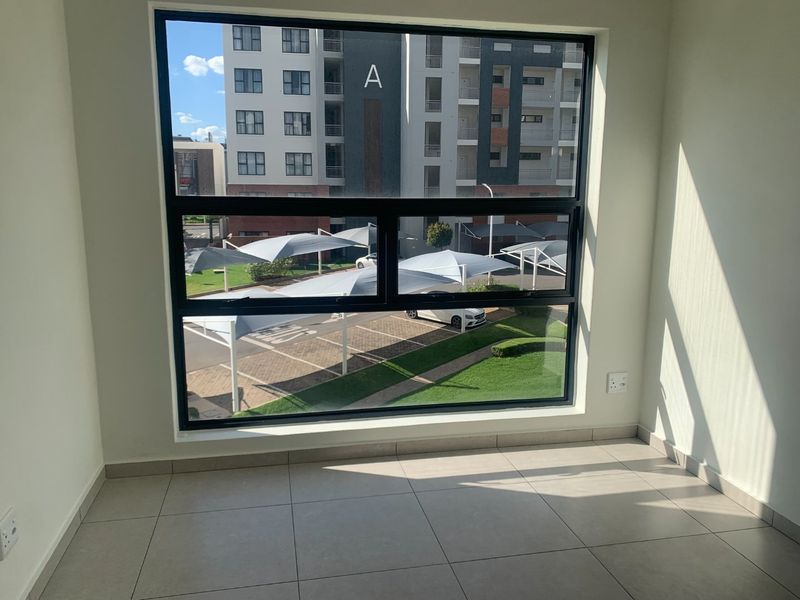 A bright, empty bedroom featuring large aluminium-framed windows with views of the complex parking area and modern tiled flooring.