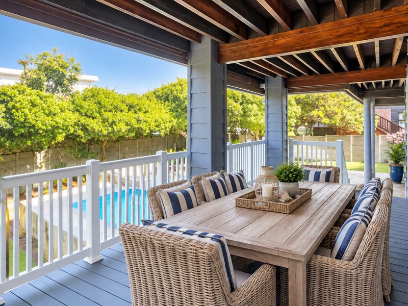 A covered outdoor patio featuring timber decking, exposed ceiling beams, and a white balustrade, overlooking a private swimming pool and garden area.