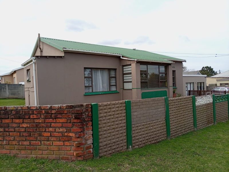 Front exterior view of a single-story house featuring a green corrugated metal roof, aluminium windows, and a combination of brick and precast concrete boundary walls.