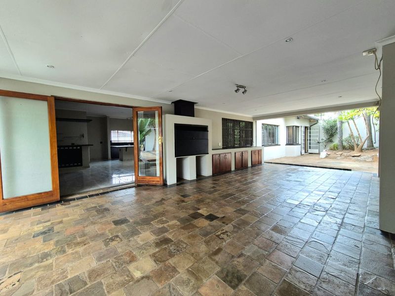 Spacious covered patio featuring stone tile flooring, a built-in braai, and wooden framed glass doors leading into the main living area.