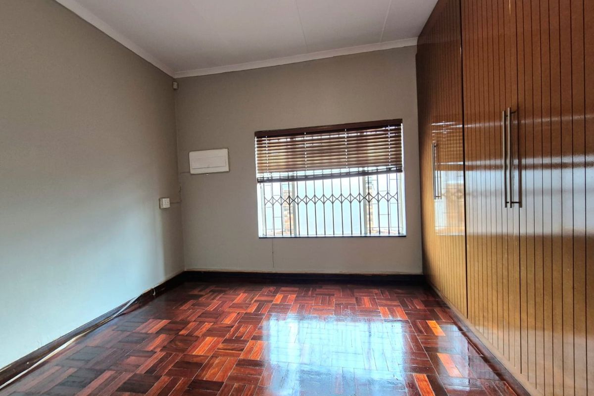 A bright, empty bedroom featuring polished parquet flooring, extensive built-in wooden cupboards, and a window fitted with security bars and venetian blinds.