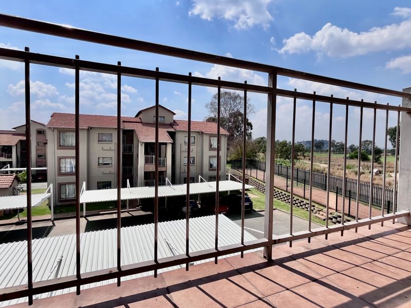 A private balcony featuring tiled flooring and a metal balustrade, offering an elevated view of the apartment complex grounds and parking area.