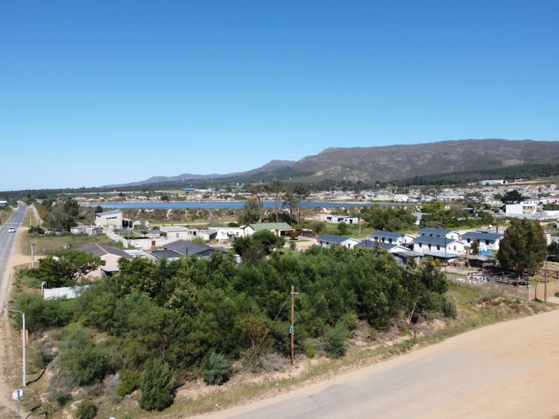 An aerial view of a vacant residential plot featuring natural vegetation, adjacent to a road with surrounding neighborhood and mountain landscape views.