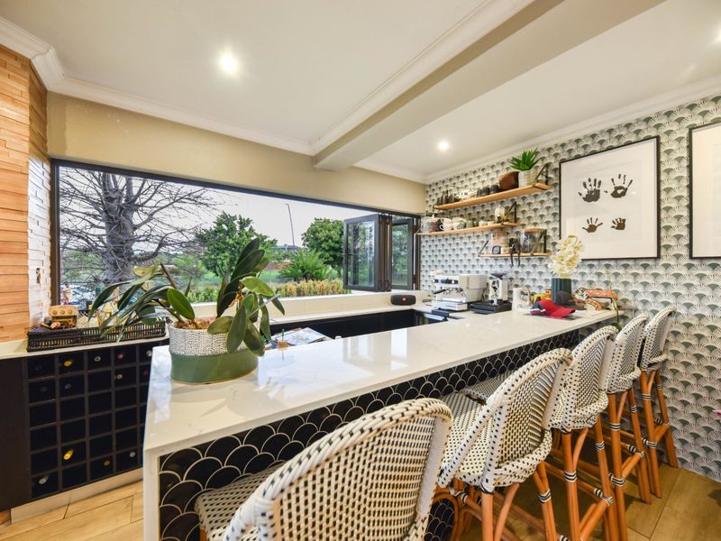 A stylish, well-appointed home bar featuring a stone-topped counter, integrated wine storage, floating shelving, and a large bi-fold window overlooking the garden.