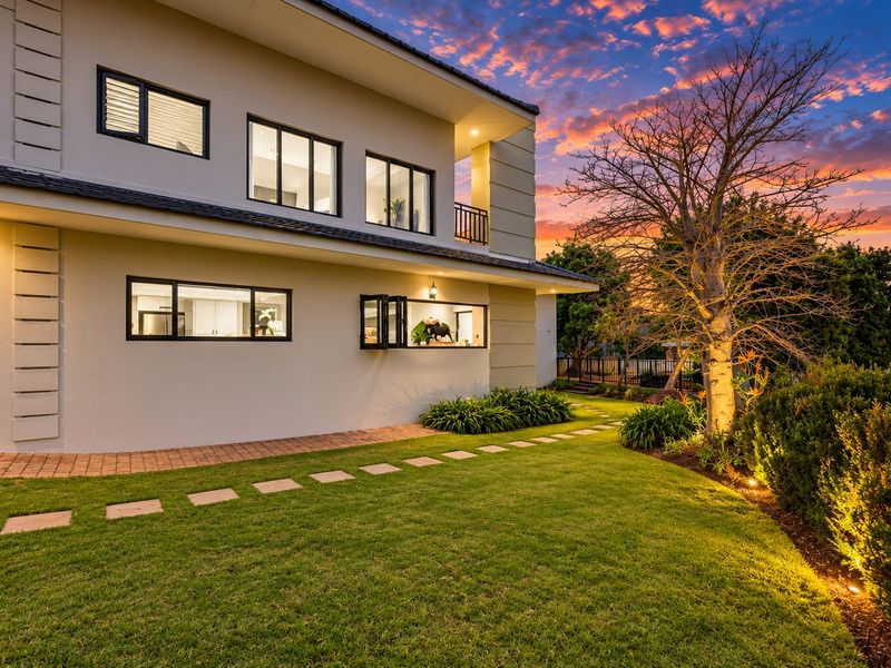 A beautifully maintained rear exterior view of a modern two-story home featuring landscaped gardens, a paver walkway, and contemporary bi-fold windows at sunset.