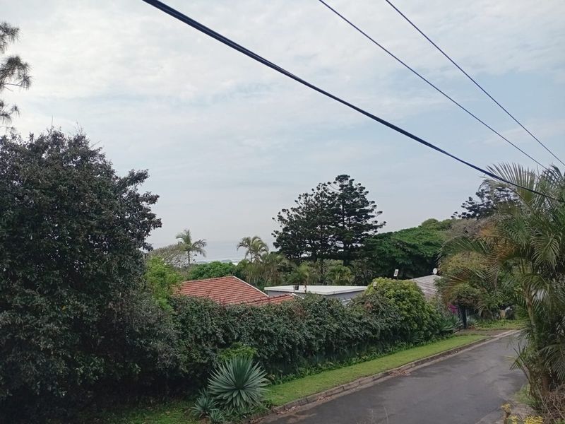 A street-level view showing the neighborhood approach, surrounding greenery, and glimpses of nearby residential rooftops.