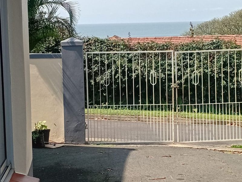 View of the property entrance featuring a metal security gate, paved driveway, and boundary wall with a glimpse of the ocean in the distance.