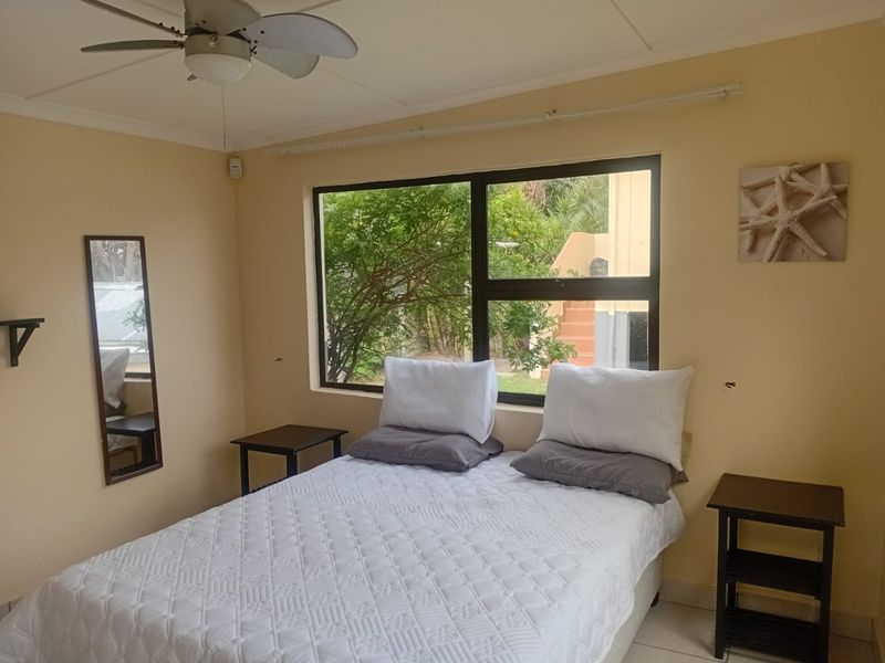 A bedroom featuring tiled flooring, a ceiling fan, and a large window with an aluminium frame, offering a view of the exterior garden area.