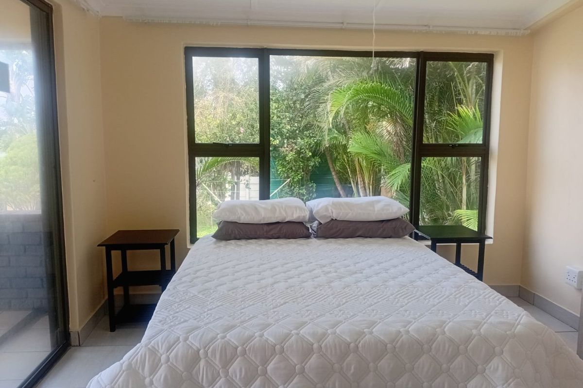 A bright bedroom featuring large aluminium-framed windows with garden views, tiled flooring, and direct access to an outdoor area via a sliding glass door.