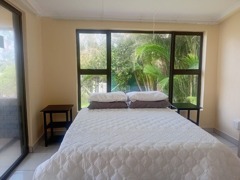 A bright bedroom featuring large aluminium-framed windows with garden views, tiled flooring, and direct access to an outdoor area via a sliding glass door.