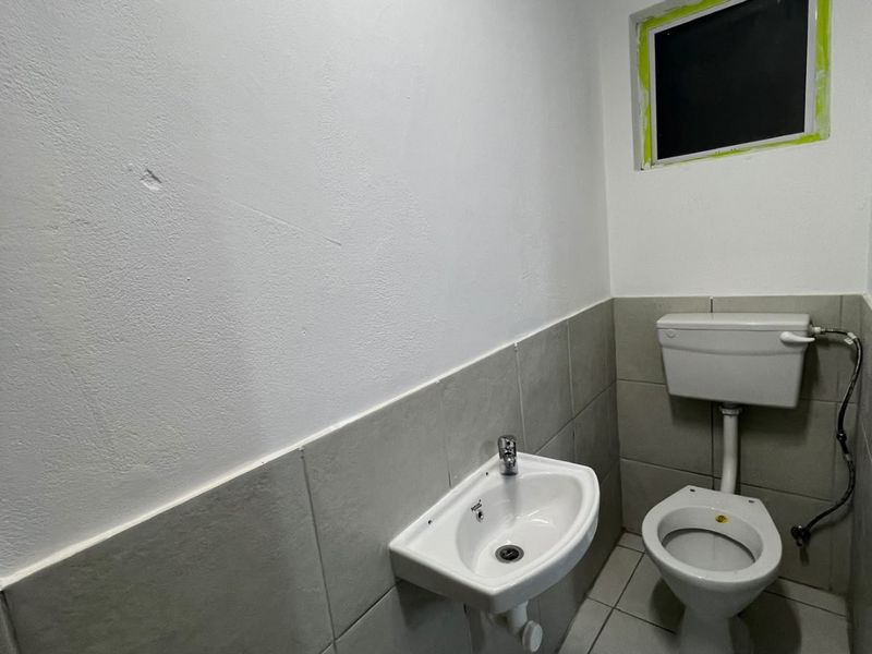 A compact, functional bathroom featuring a wall-mounted basin, toilet, and neutral tiled finishes, currently awaiting final trim completion around the window.