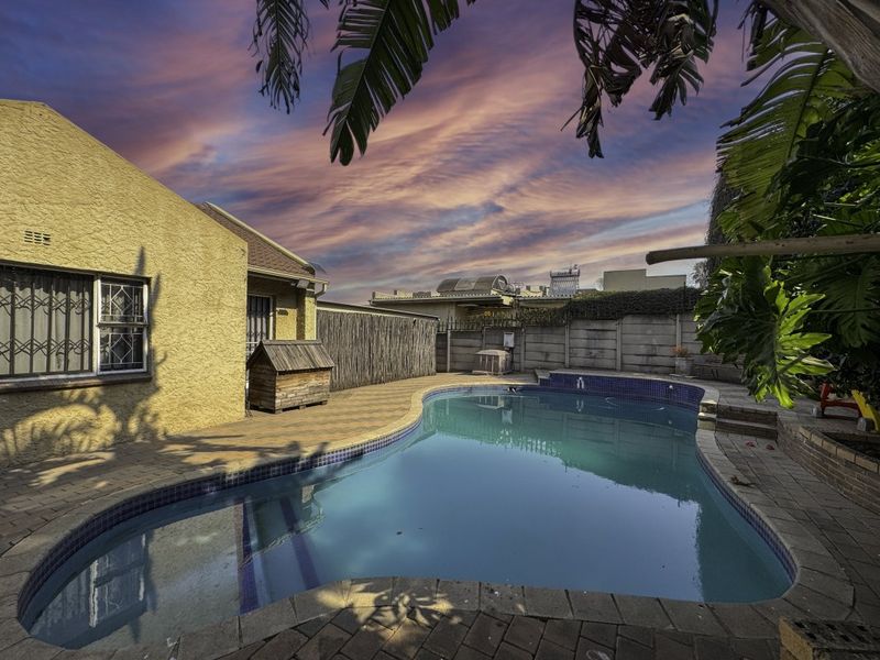Outdoor pool area featuring a paved patio, swimming pool, and perimeter walling, set against a sunset sky.