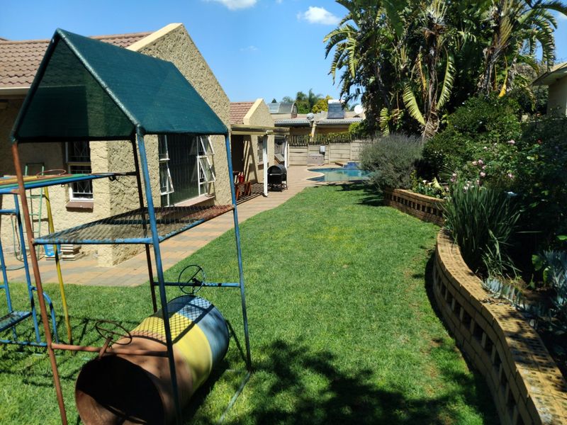 Rear garden view featuring a paved patio area, swimming pool, and established landscaping with a retaining wall and tiled roof house exterior.