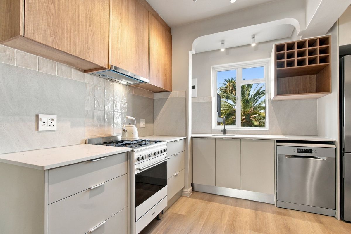 Modern kitchen featuring light wood cabinetry, stone countertops, gas stove, extractor fan, and integrated dishwasher with a bright window view.