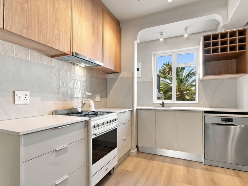 Modern kitchen featuring light wood cabinetry, stone countertops, gas stove, extractor fan, and integrated dishwasher with a bright window view.