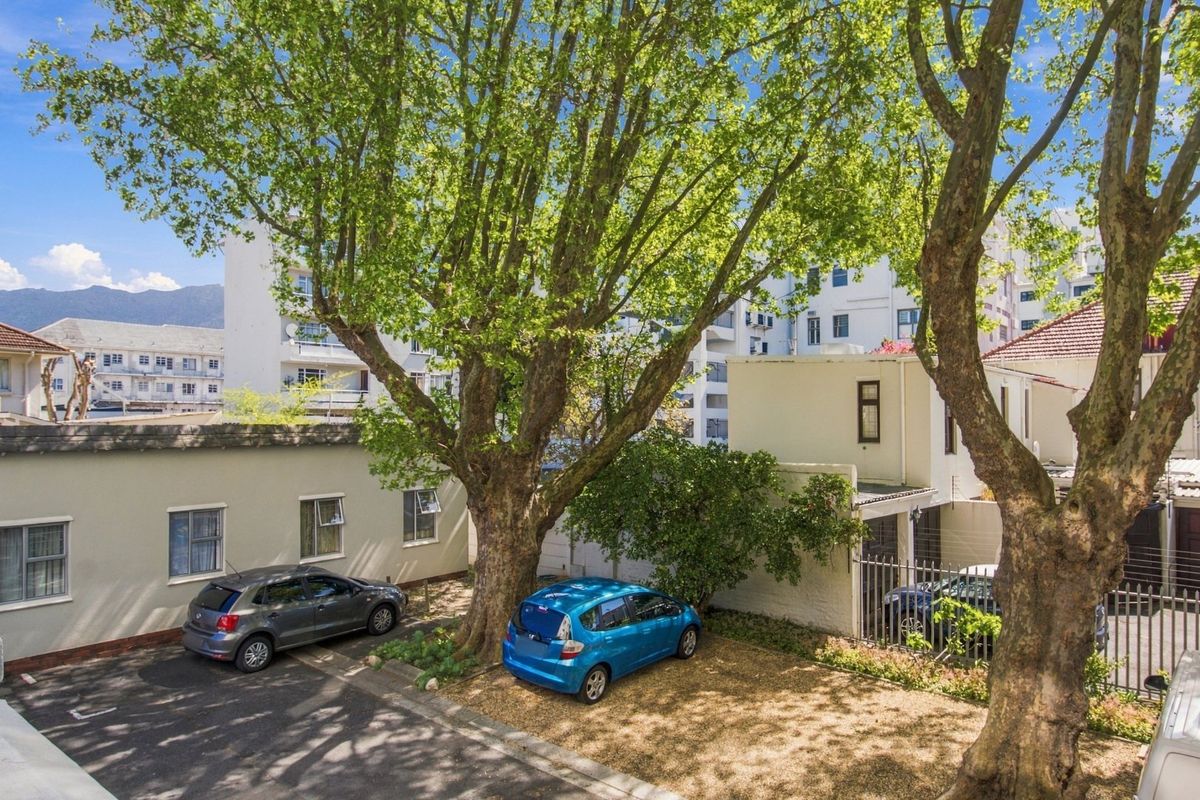 Rear exterior view of the apartment complex featuring a paved driveway, gravel parking area, and mature trees providing shade.