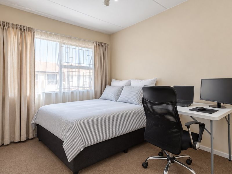 A well-maintained bedroom featuring neutral wall tones, carpeted flooring, and large windows fitted with security bars and curtain tracks.