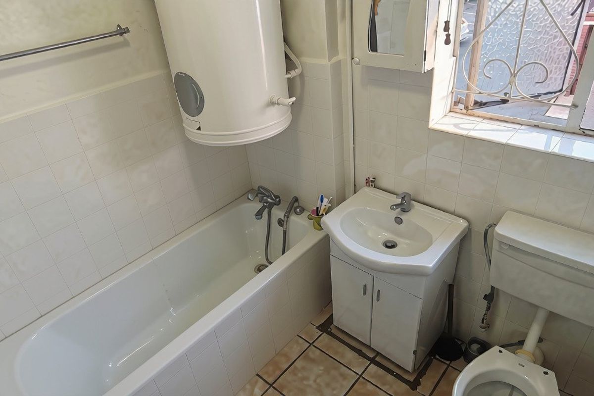A compact bathroom featuring a bathtub, wall-mounted water heater, vanity basin, and toilet, with tiled finishes throughout.