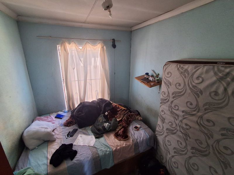 A small bedroom featuring a window with a curtain rod and a wall-mounted shelf, requiring cosmetic updates to the walls and finishes.