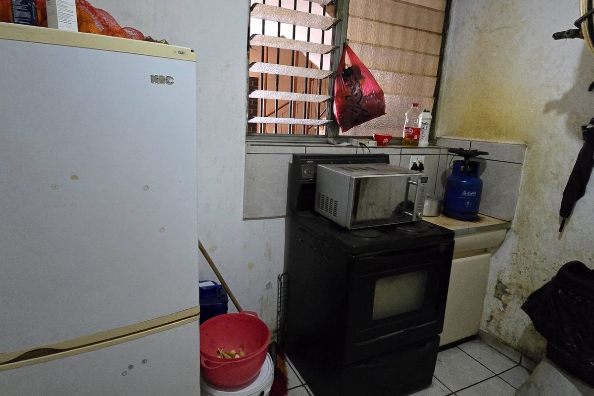 A small, dated kitchen area featuring a tiled backsplash, louvered window, and freestanding oven, currently showing significant signs of wear and wall deterioration.