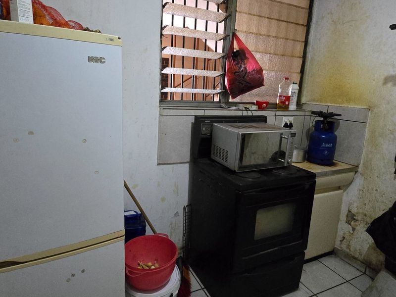 A small, dated kitchen area featuring a tiled backsplash, louvered window, and freestanding oven, currently showing significant signs of wear and wall deterioration.