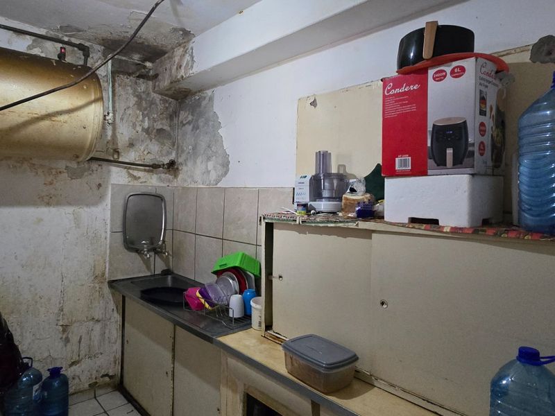 A small kitchen area featuring a stainless steel sink, wall-mounted geyser, and built-in cabinetry, currently showing significant signs of wall deterioration and wear.