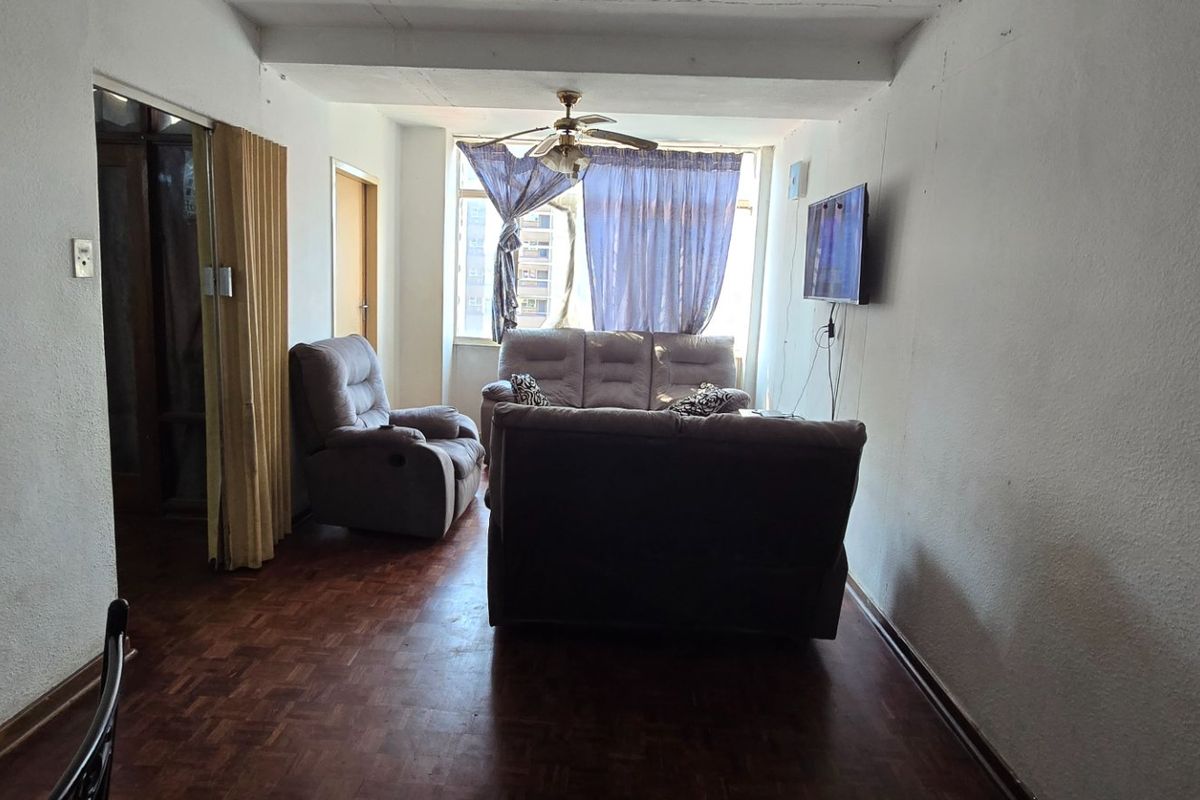 A living room featuring parquet flooring, a ceiling fan, and a folding door, providing a functional space with potential for cosmetic updates.