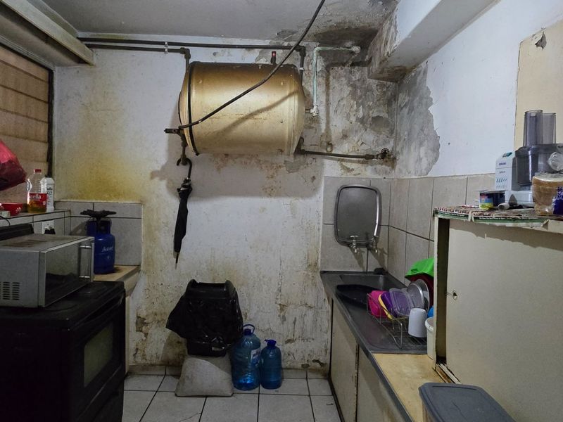 A dated kitchen area featuring a stainless steel sink, tiled backsplash, and wall-mounted water heater, currently requiring significant renovation and wall repairs.